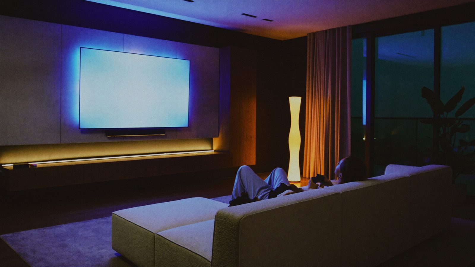 A modern open-plan lounge at night, the wall behind the screen glowing softly with ambient light.