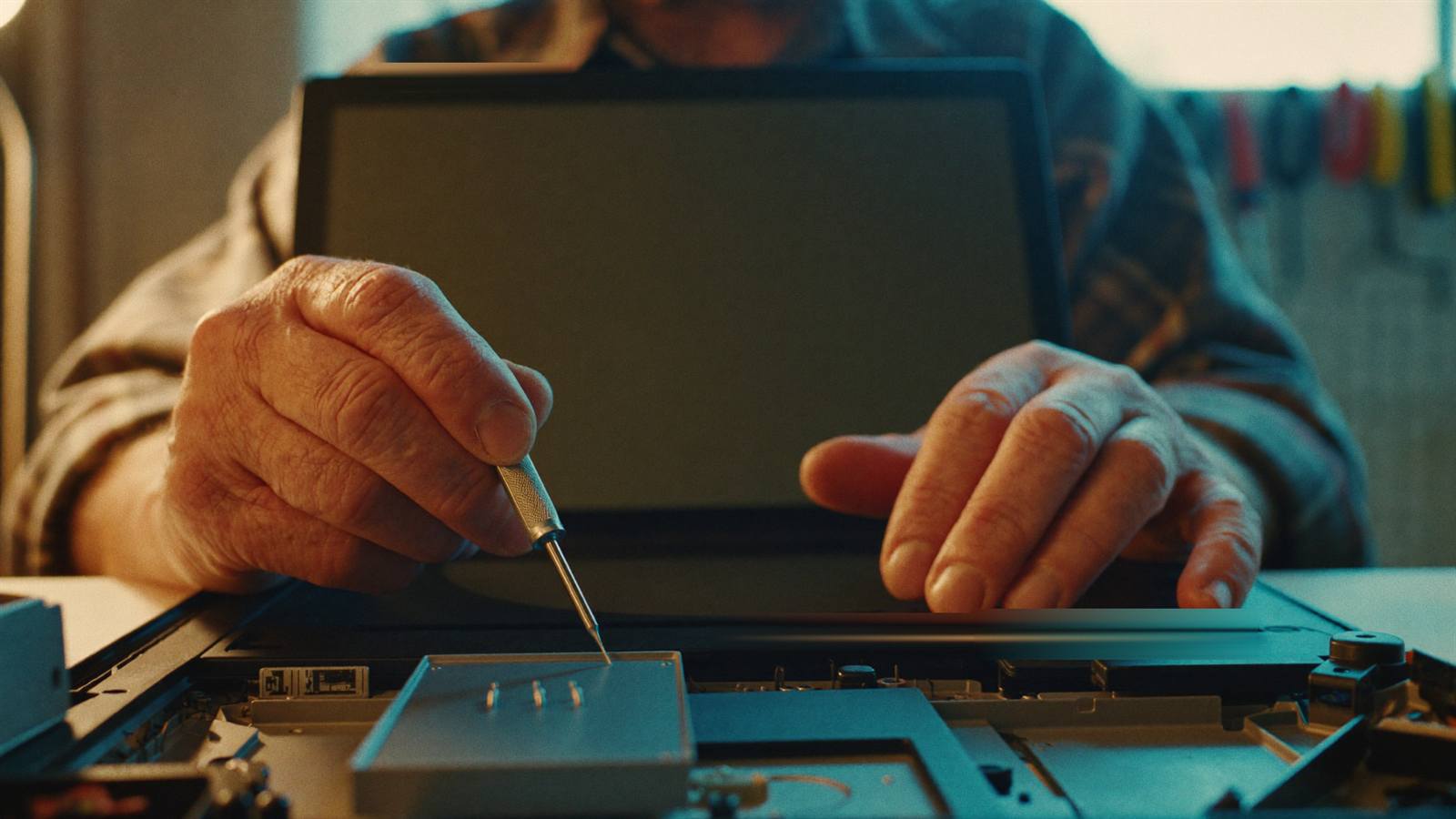 Weathered hands with a precision screwdriver over an open laptop at a tidy workbench, tools soft-focus on the pegboard behind