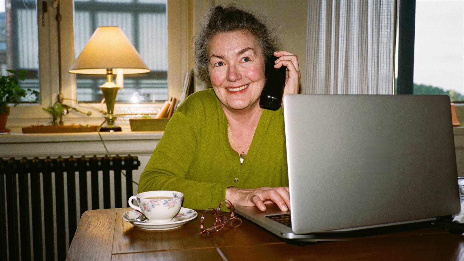 A woman at her kitchen table smiling with relief on a phone call, laptop closed beside her tea — the problem talked through and sorted
