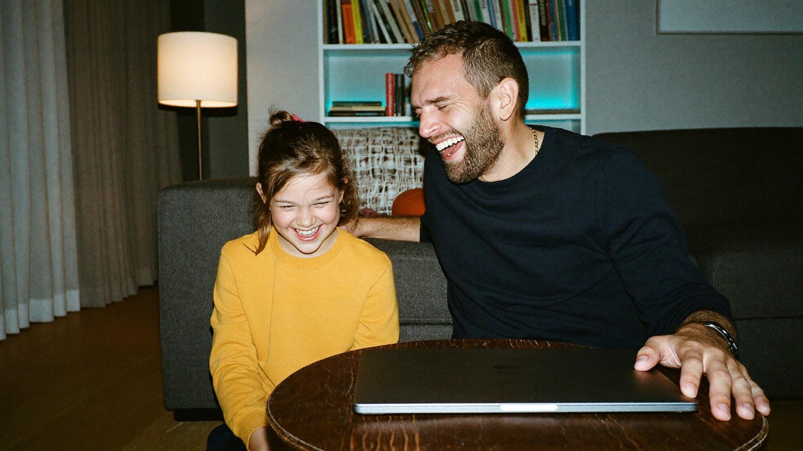 A father and daughter relaxed on the sofa in the evening, laughing, the laptop closed on the ottoman — everything working again