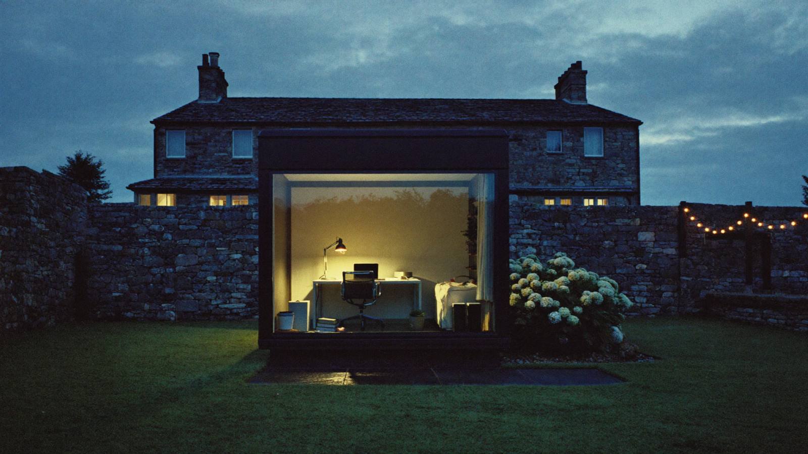 A garden office pod glowing warm at blue hour behind a stone house, with lawn and a slate path in between