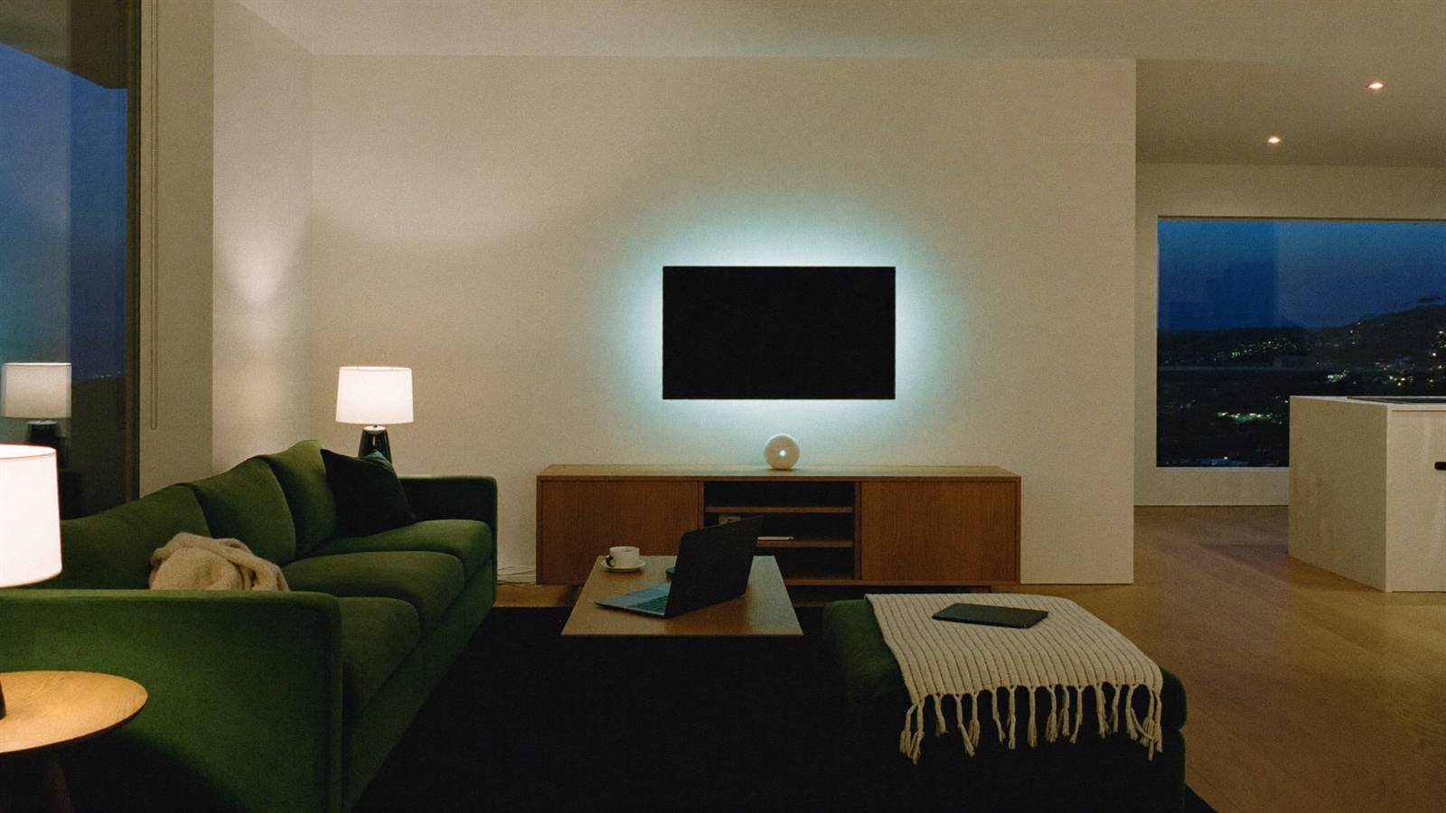 A calm modern open-plan living space at dusk, lamps glowing warm, a laptop and tablet resting closed